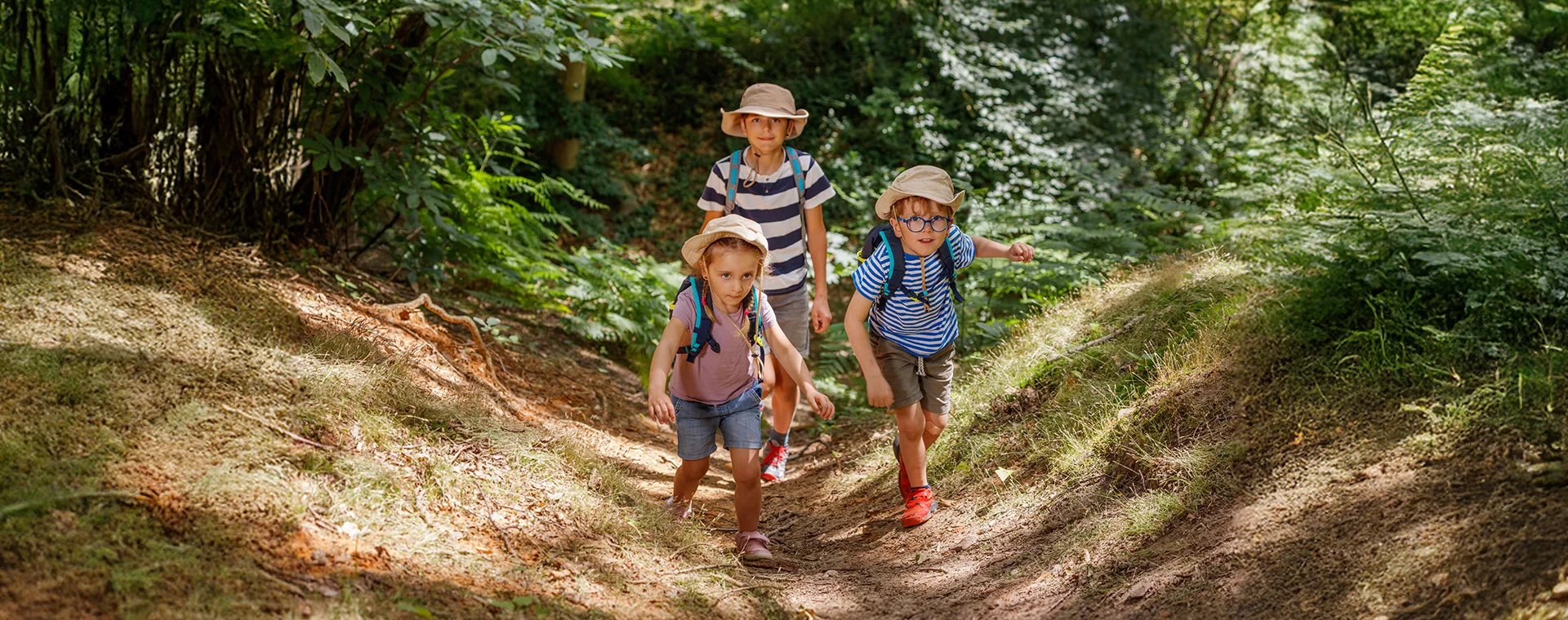 Kids Exploring Forest Trail With Backpacks And Hats On Sunny Day Kids Exploring Forest Trail With Backpacks And Hats On Sunny Day