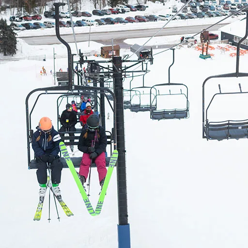 Skiers riding chair lift up snowy hill with M45 in background at Marquette Mountain Resort